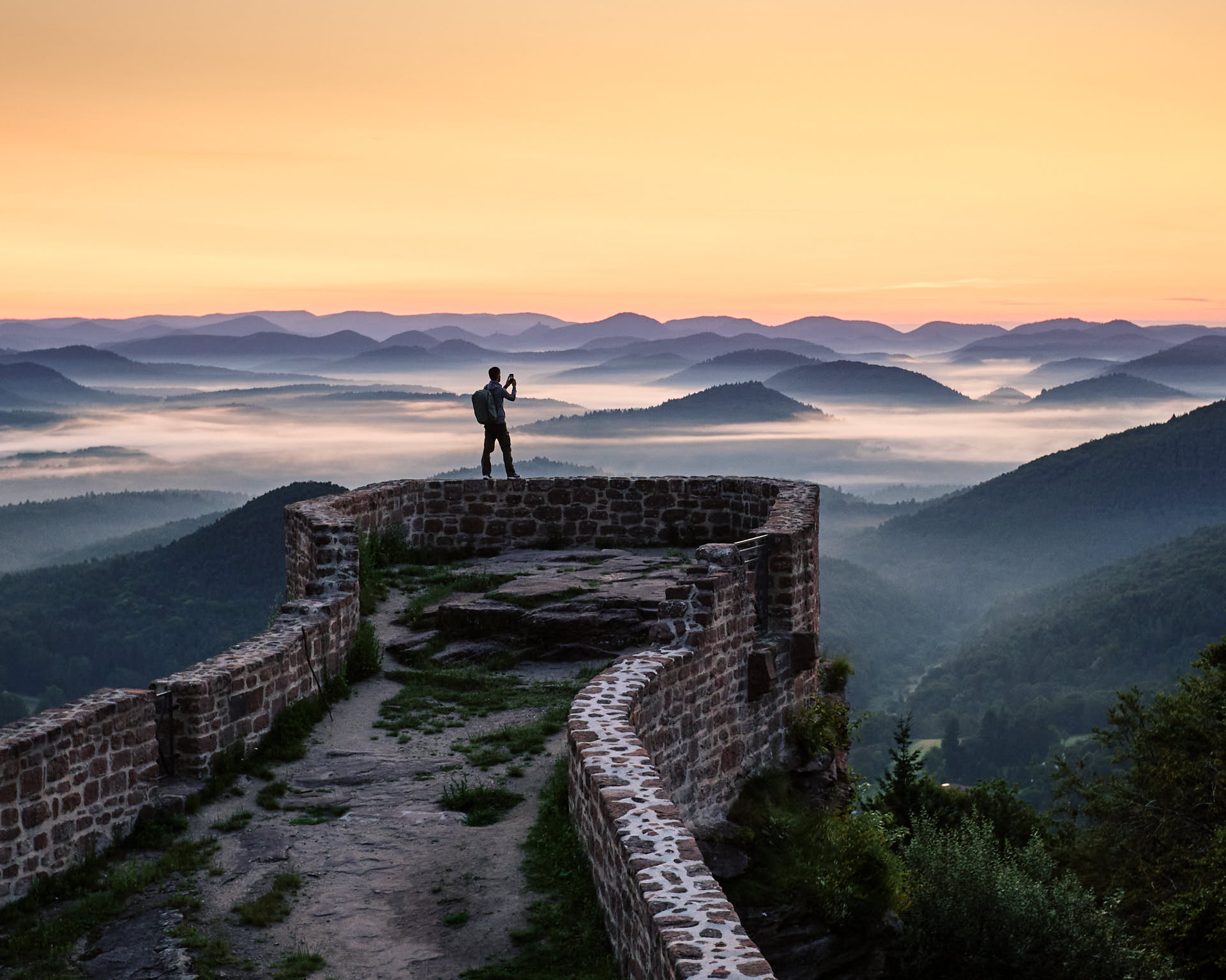 Eine Person steht als Silhouette auf der Mauer von der Wegelnburg. Im Hintergrund Bergketten und Nebel in den Tälern. Das Bild ist kurz vor Sonnenaufgang aufgenommen.